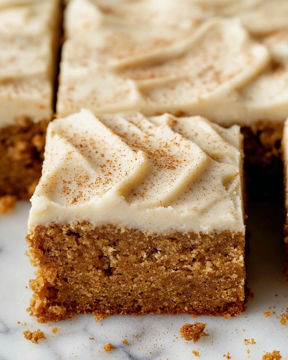 The image shows a close-up of a square piece of cake with two clear layers. The bottom layer is a thick, moist cake with a textured, crumbly brown surface. Above it is a smooth, thick layer of creamy white frosting, spread evenly with subtle swirls and a light dusting of brown spice powder on top. The cake is cut into square pieces sitting on a white marbled surface with small crumb bits scattered around. photo taken with an iphone --ar 4:5 --v 7