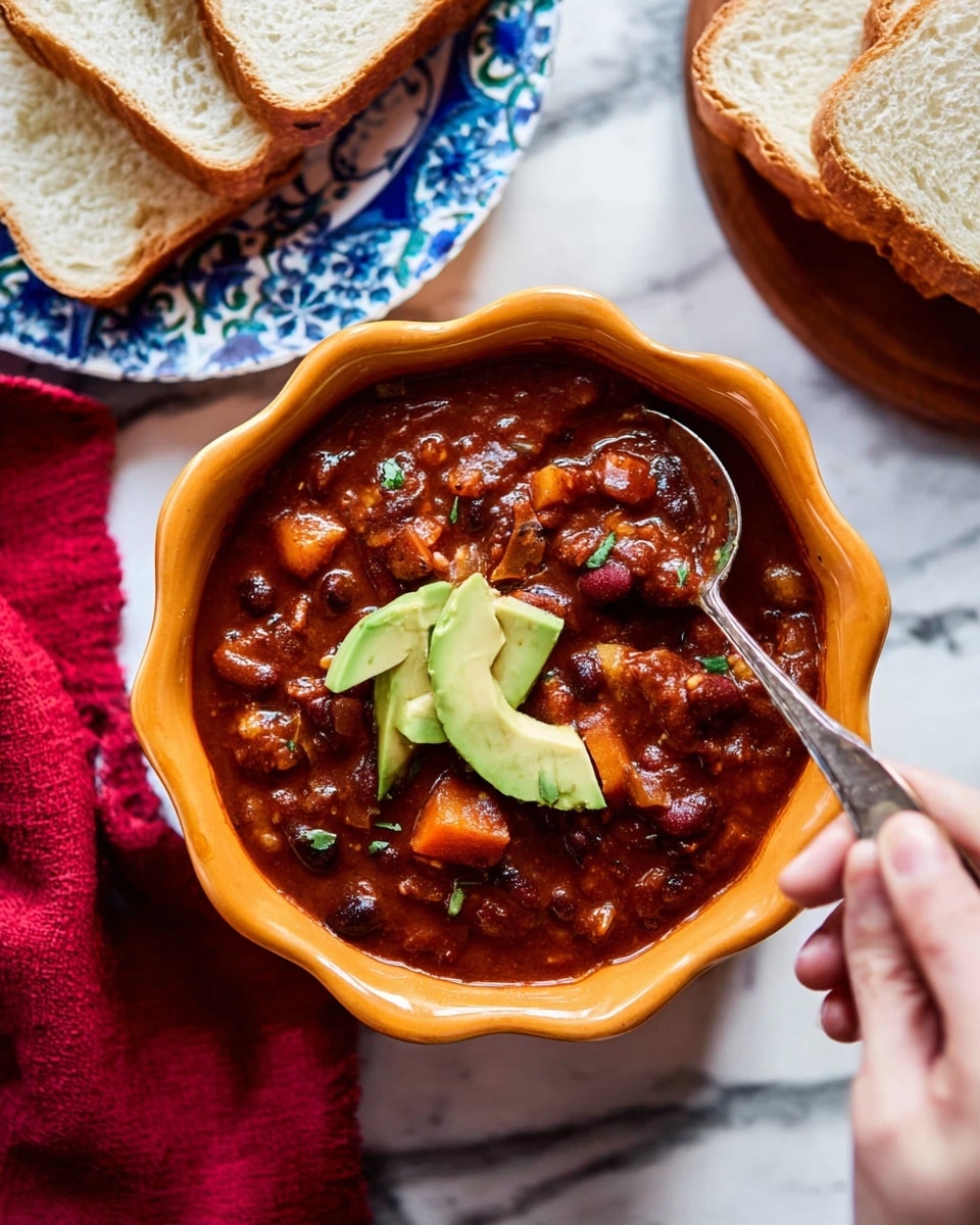 The image shows a bowl of thick chili with visible chunks of vegetables and beans in a rich, dark reddish-brown sauce, filled to the top. Two slices of fresh green avocado sit on the chili’s surface as a garnish. The bowl is scalloped and orange-colored, resting on a white marbled surface. A woman's hand is holding a spoon dipping into the chili from the right side. In the background, there is a blue patterned plate with thick slices of white bread, and a red cloth napkin is partially visible on the left side. Photo taken with an iphone --ar 4:5 --v 7