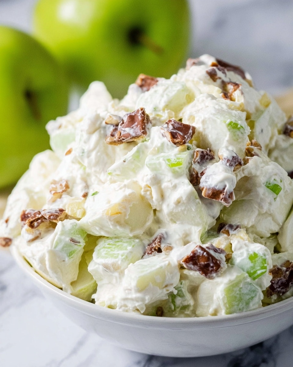 A close-up view of a creamy white salad served in a white bowl, showing chunks of light green apple pieces layered with creamy white whipped topping mixed with chunky brown candy or chocolate pieces, giving the salad a fluffy texture with scattered bits of green celery or similar vegetable throughout. The background shows a blurred green apple and the bowl sits on a surface with a white marbled texture. Photo taken with an iphone --ar 4:5 --v 7