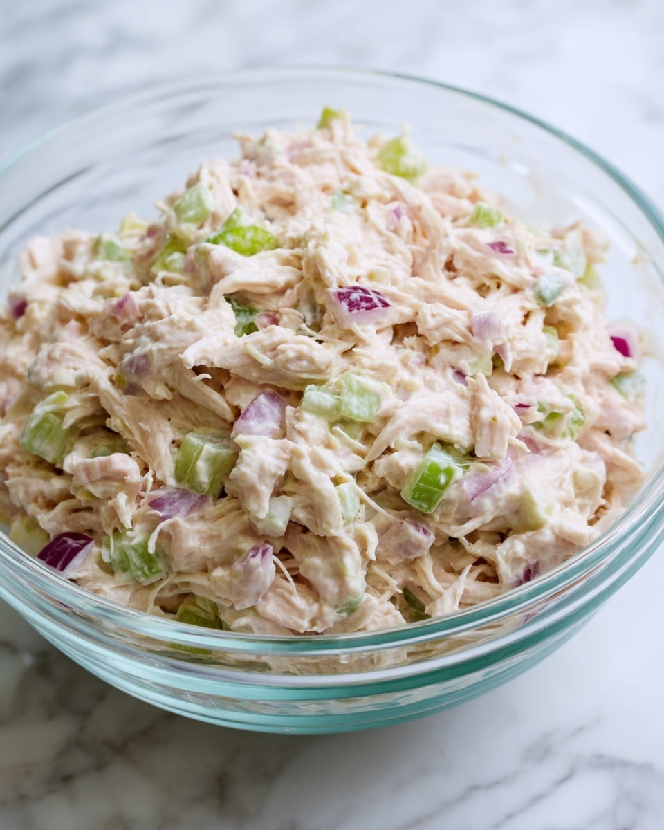 A close-up view of a clear glass bowl filled with a creamy shredded chicken salad. The salad has a light pinkish-white creamy texture with fine shreds of chicken mixed throughout. Small pieces of green celery and bits of purple-red onion are scattered evenly inside, adding color and texture. The creamy mixture looks thick but soft, with a slightly chunky feel due to the vegetables and chicken. The bowl is placed on a white marbled surface that contrasts gently with the salad colors, enhancing its fresh appearance. photo taken with an iphone --ar 4:5 --v 7