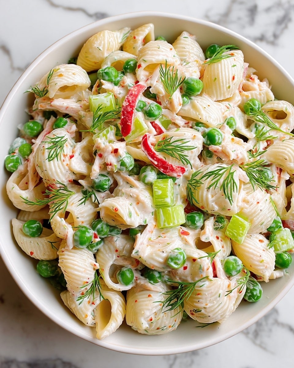The image shows a close-up of a creamy pasta salad in a white bowl on a white marbled surface. The dish has large shell-shaped pasta, which is a pale yellow, mixed with bright green peas and light green chopped celery. There are also pieces of red bell pepper and shredded white chicken mixed throughout. Fresh green dill is sprinkled on the top, adding a light and fresh look. The creamy dressing coats all the ingredients, giving a smooth and slightly glossy texture to the pasta and vegetables. photo taken with an iphone --ar 4:5 --v 7