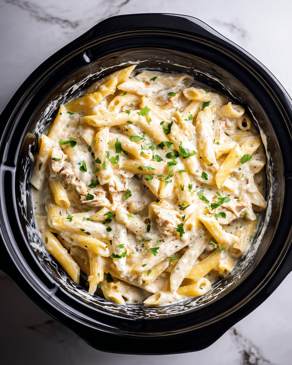 The image shows a black slow cooker bowl filled with creamy pasta. The pasta is penne, coated in a thick white sauce that looks smooth and rich. There are small pieces of chicken mixed throughout, and tiny green herbs sprinkled on top, adding a pop of color. The edges of the bowl have some sauce smudges, showing the dish was stirred. The background has a white marbled texture surface. photo taken with an iphone --ar 4:5 --v 7