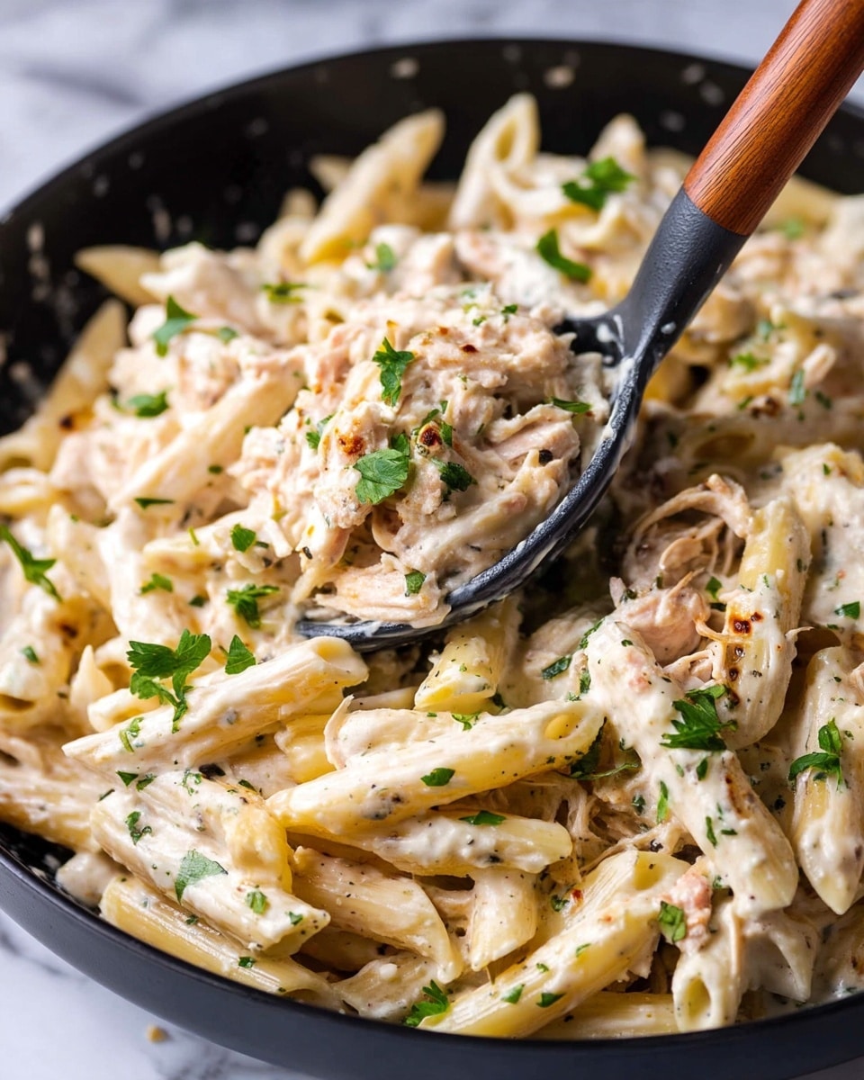 The image shows a close-up of creamy white penne pasta mixed with a thick, rich white sauce that has visible herbs and small green flecks scattered throughout. The pasta pieces are coated evenly in the sauce, and there are small bits of light pink shredded chicken mixed in. Fresh green parsley pieces are sprinkled on top for color contrast. The pasta is being scooped with a black spoon that has a brown wooden handle. The background is a black dish with some sauce smudges on the sides, sitting on a white marbled surface. Photo taken with an iphone --ar 4:5 --v 7