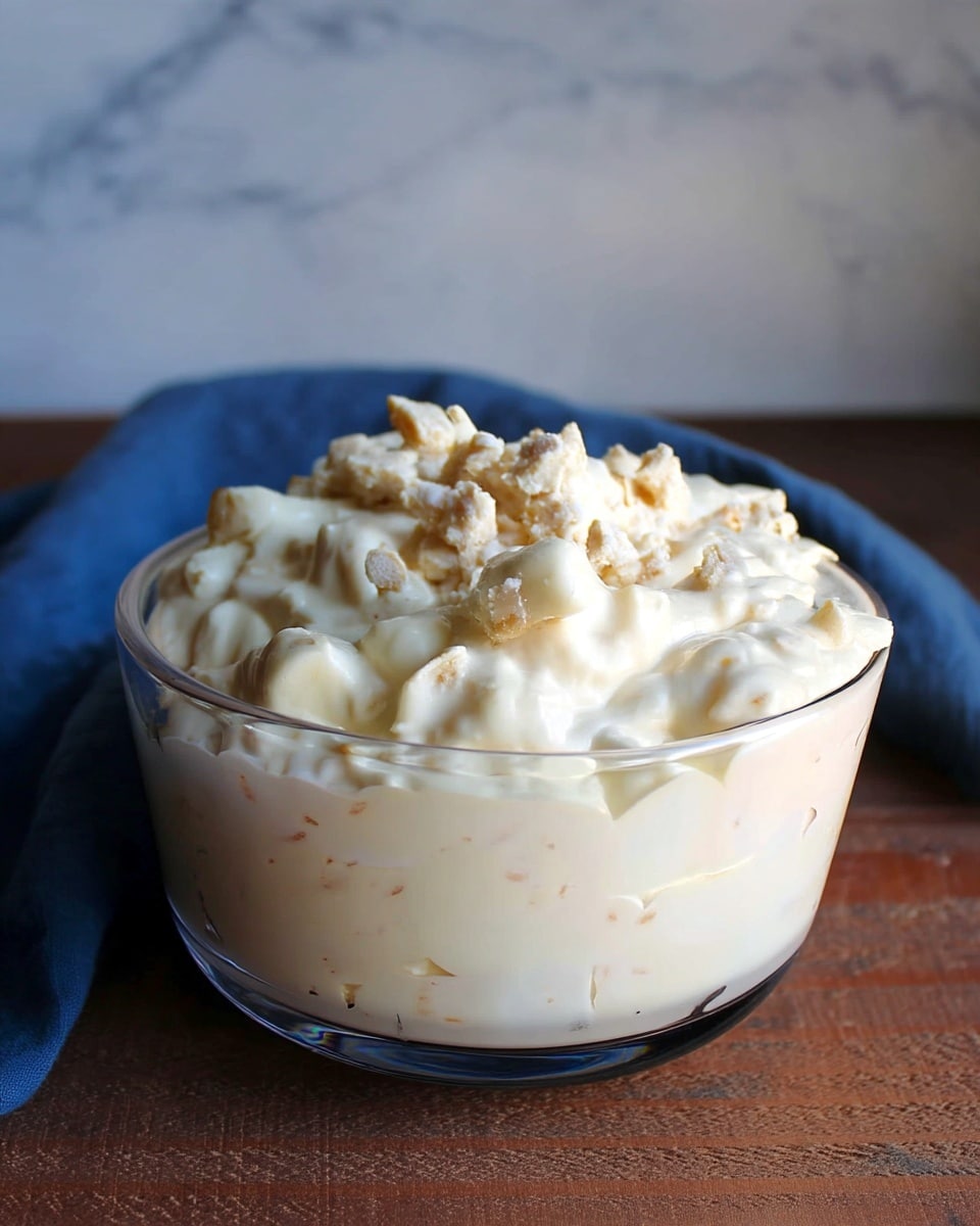A clear glass bowl filled with a creamy dish that has two visible layers: the bottom layer is smooth and pale cream-colored, while the top layer is thicker and chunky with light beige pieces coated in the same creamy texture. The bowl is placed on a dark wooden surface with a blue cloth in the background, all set against a white marbled texture. photo taken with an iphone --ar 4:5 --v 7
