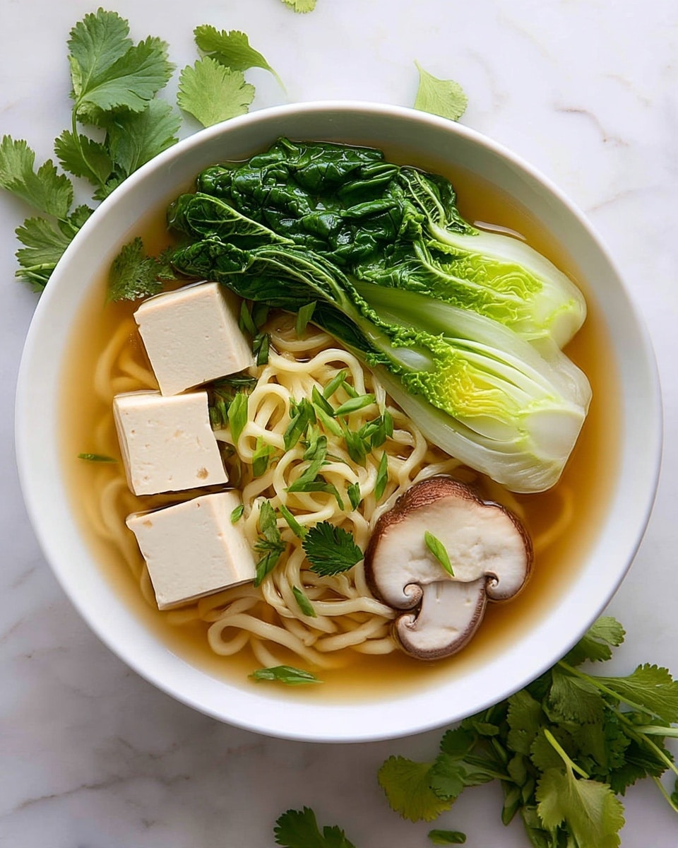 A white bowl holds a clear broth filled with three large cubes of pale tofu on the left side, bright green bok choy leaves with a light green stem at the top right, and two slices of light brown shiitake mushroom with a creamy white center on the right edge. The center and bottom layers show curly, pale yellow noodles, lightly sprinkled with chopped green onions. Some fresh cilantro leaves float on top, adding bright green color around the tofu and noodles. The bowl is placed on a white marbled surface, surrounded by scattered cilantro leaves. Photo taken with an iphone --ar 4:5 --v 7