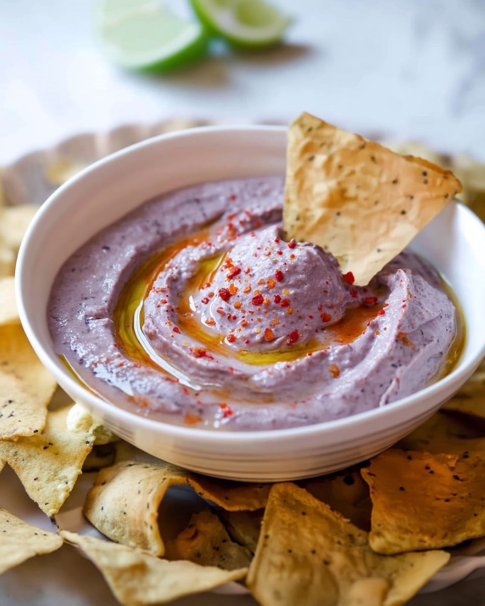 A white bowl filled with a creamy purple dip that has a smooth, slightly thick texture and swirled surface. The dip is garnished with a thin drizzle of golden oil and tiny red spice flecks scattered on top. One large triangular corn chip with speckles is resting inside the bowl, partially dipped, while more chips surround the bowl at the base, all placed on a white marbled surface. Photo taken with an iphone --ar 4:5 --v 7
