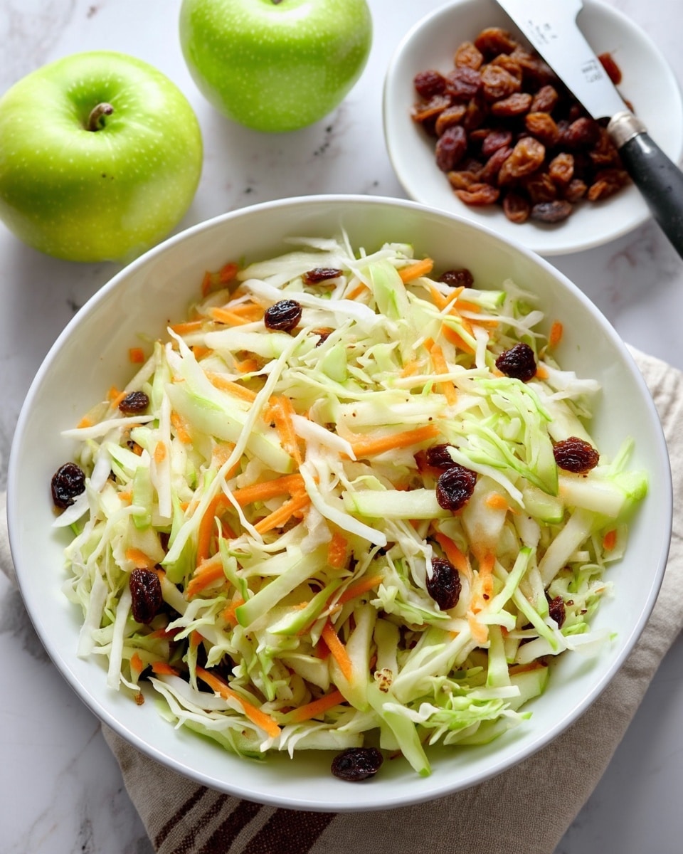 The image shows a white bowl filled with a mix of shredded pale green cabbage, thin orange carrot strips, and small dark brown raisins scattered throughout, creating a fresh and colorful salad mix. The bowl sits on a white marbled surface, with two whole green apples and one half apple placed nearby. A white dish with more raisins and a knife with a black handle are also visible in the background. Photo taken with an iphone --ar 4:5 --v 7