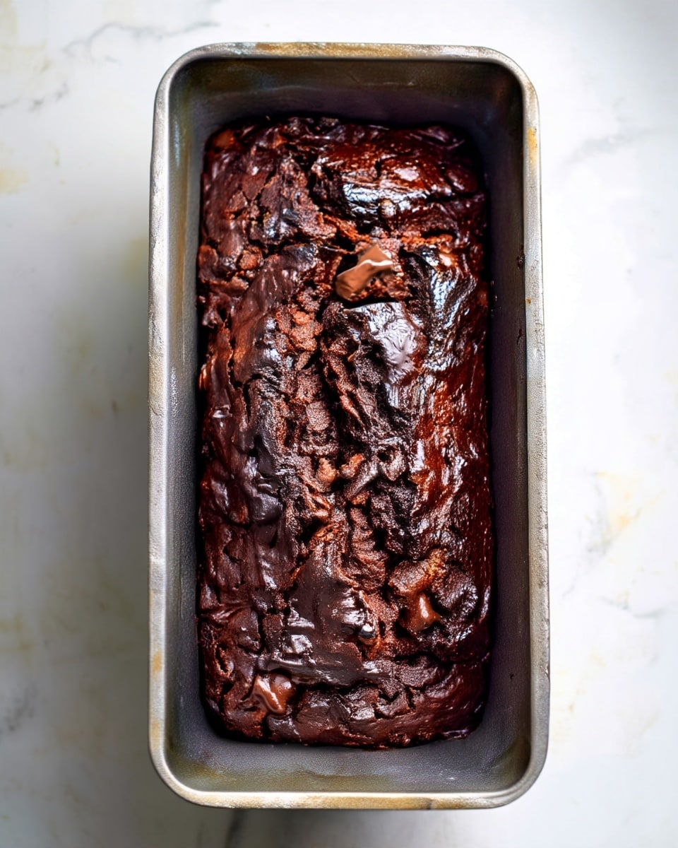 A loaf of dark chocolate bread with a rich, textured surface, filled with small chunks of melted chocolate, is placed inside a rectangular metal baking pan. The bread shows deep cracks and uneven rises on top, revealing its gooey chocolate interior. The metal pan looks slightly worn and is resting on a white marbled surface. photo taken with an iphone --ar 4:5 --v 7