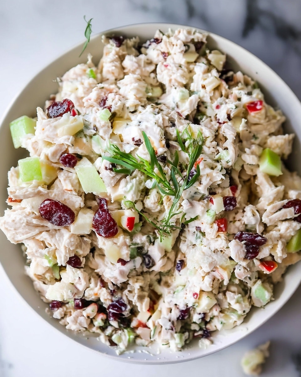 A close-up view of a white bowl filled with creamy chicken salad that has a thick, mixed texture showing finely chopped white chicken pieces combined with small chunks of light green celery, bits of red apple, dark red dried cranberries, and light brown nuts, all coated well in creamy white dressing. The salad is garnished on top with a few fresh green sprigs of herbs. The bowl is placed on a white marbled surface. photo taken with an iphone --ar 4:5 --v 7